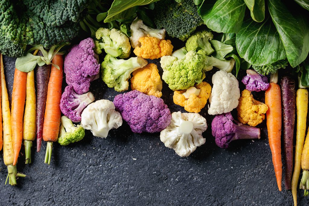 An array of colorful carrots, broccoli, purple broccoli, cauliflower, and lettuce on a black background