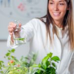 smiling female botanist working in a lab full of plants holding a vial ful of clear liquid. 