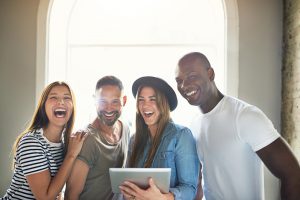 four friends standing in front of a large window smiling and laughing into the camera