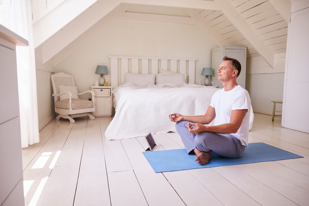 Middle aged man in an all white bedroom meditatiing peacefully facing the window on a blue yoga matt.