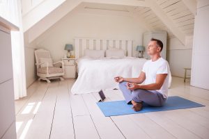 relaxed man sitting legs crossed on a blue yoga mat in his room meditating 