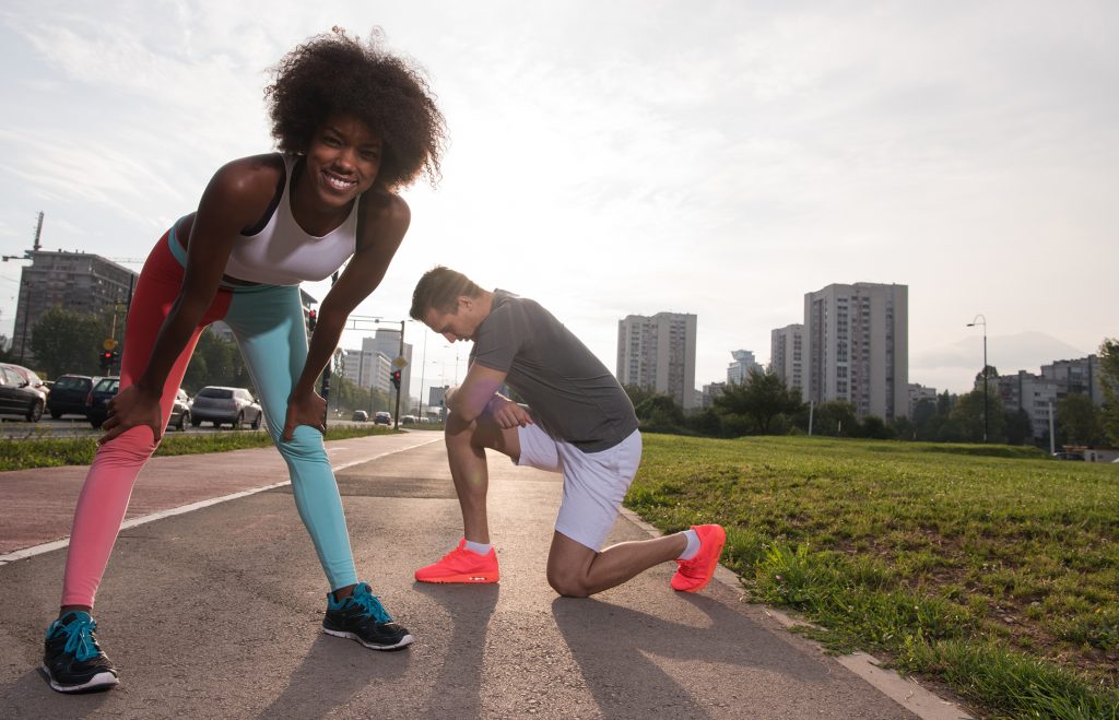 Young woman in running gear bending down smiing into the camera as her male partner is kneeing on the asphalt resting. City landsape in teh background