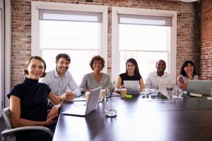 Group of happy smiling coworkers sitting around a large table with computers out  in a large meeting room