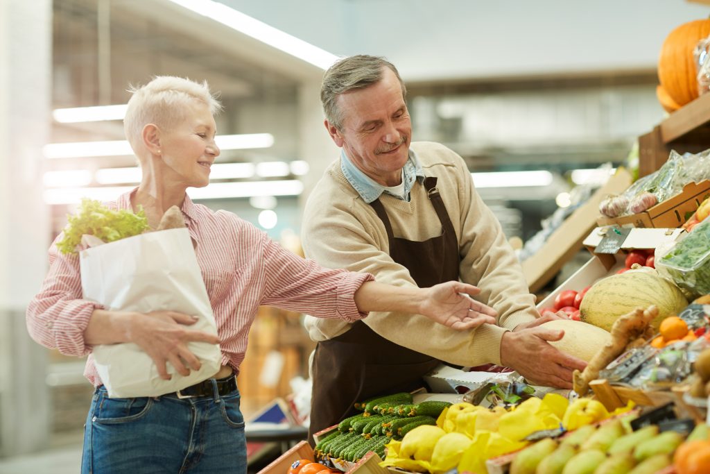 An elder male grocer helping a older fit woman who is holding a bag full of groceries to a watermelon that is on display with other fruits and vegetables