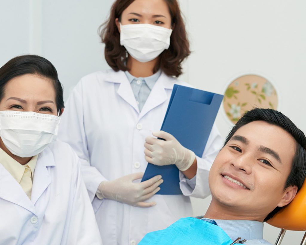 female dentist and dental hygenist holding a blue clipboard are  smiling next to a male patient wearing a blue cloth bib