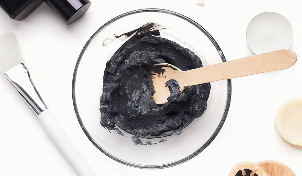 white bowl filled with activated charcoal stiired with a wooden spoon into a paste. Next to the bowl is a makeup brush