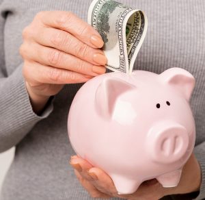 woman wearing a grey sweater putting a hundred dollar bill in a pink piggy bank