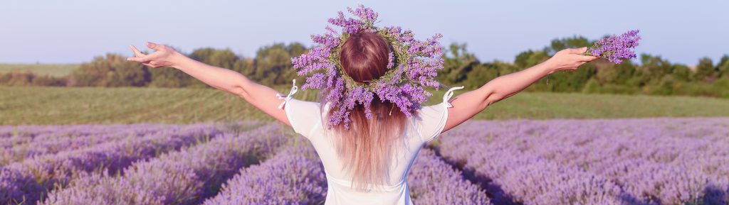 young woman dressed in white wearing a crown of lavander plants arms stretched in front of a lavander field