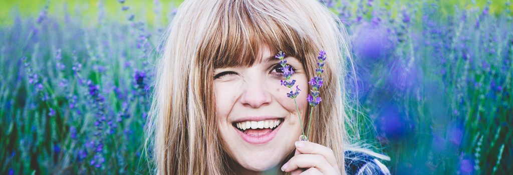 Young woman in a lavader field smiling as she holds up s lavander stalk to her eye.