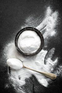 Wooden bowl and spoon filled with baking soda sitting on a table that has baking soda ll over it. 