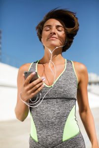 Young woman in grey work out clothes smiling with hey eyes closed and heaphones in listening to music. 