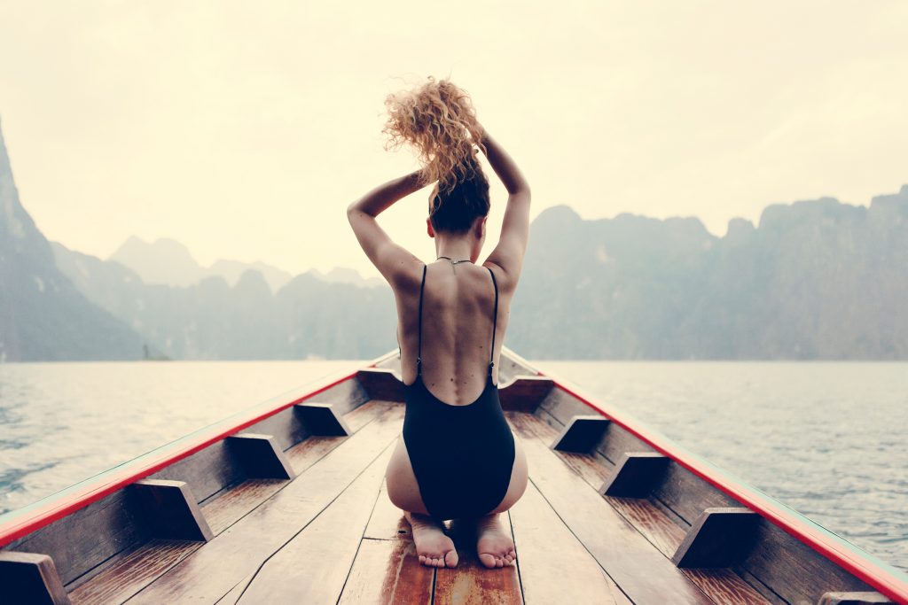 young fit woman in a black backless bathing suit putting her hair in a ponytail whole kneeling on the bow of a boat that is in the ocean with mountains in the background