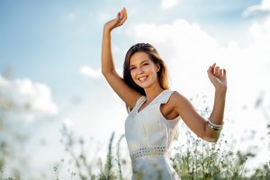 Smiling young woman in white, outside in a field stretching her arms up to the sky that is full of large fluffly clouds