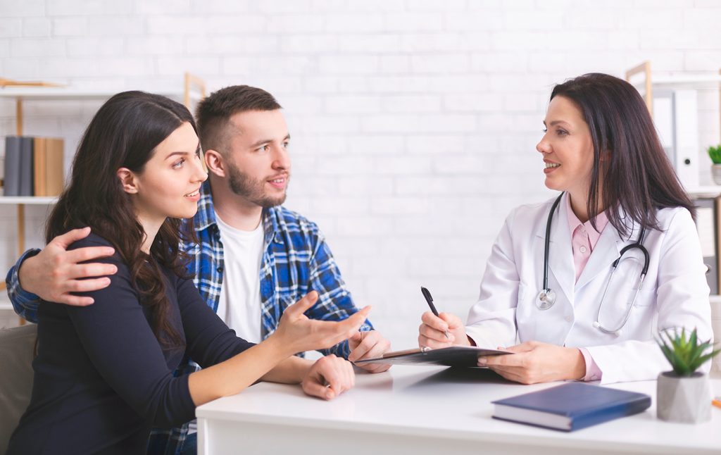 young couple sitting at a desk  in a doctor's office discussing options with the female doctor who is taking notes on a clipboard. 