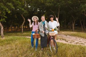 three woman standing next to a groove of trees. The young woman on the left is holding a basket a ful of fruits and vegetables waving to the camera. The woman in the middle has a bike with a basket also filled with fruits and veggies. The womamn on the right is smiling and waving to the camera