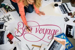 young woman in a pink sweater and blue jeans  working on a large pink BON VOYAGE sign surrounded by pictures, books, and other signs