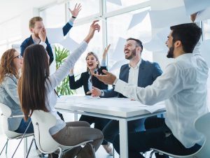 smiling laughing coworkers sitting around a square desk with their hands raised